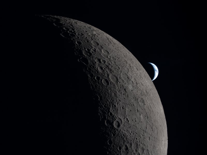 The Moon lit from one side, its cratered limb in raking sunlight, with a crescent Earth beyond it.